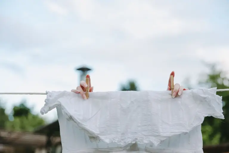 Close-up of hands pinning a white blouse on a clothesline outside on a sunny day.