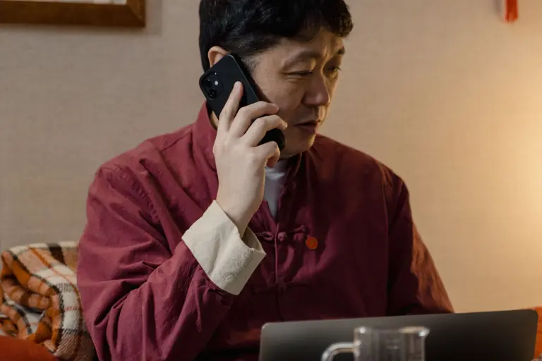 An adult man in traditional attire talks on the phone while working on a laptop indoors.