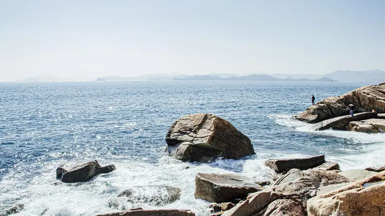 A serene coastal landscape with rocky shores, waves crashing, and distant hazy mountains.