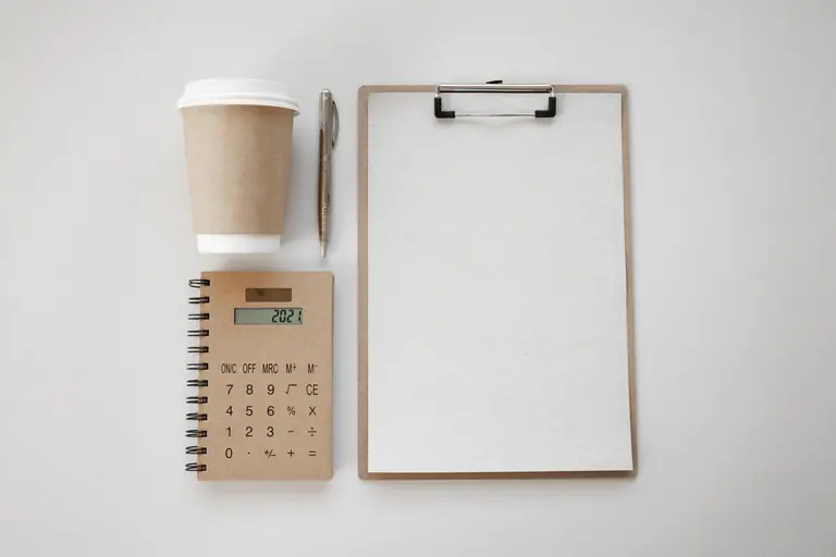 Flat lay of a minimalist office workspace with coffee cup, clipboard, pen, and notebook calculator.