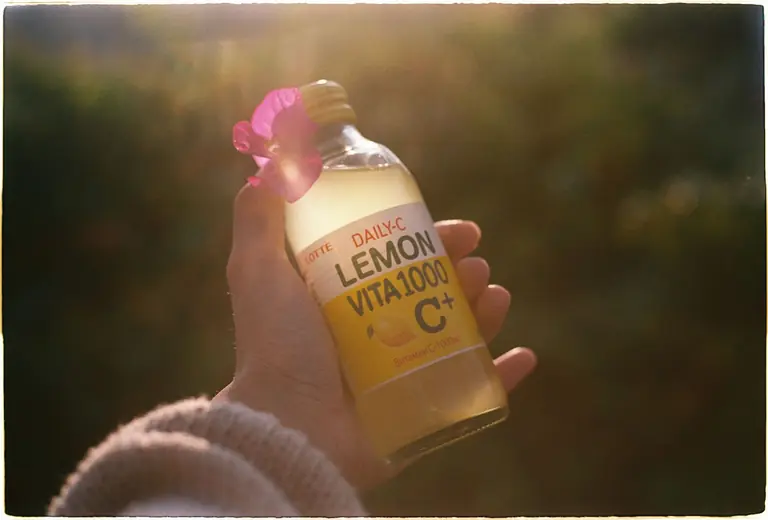 A hand holding a lemon-flavored vitamin drink bottle outdoors in sunlight.