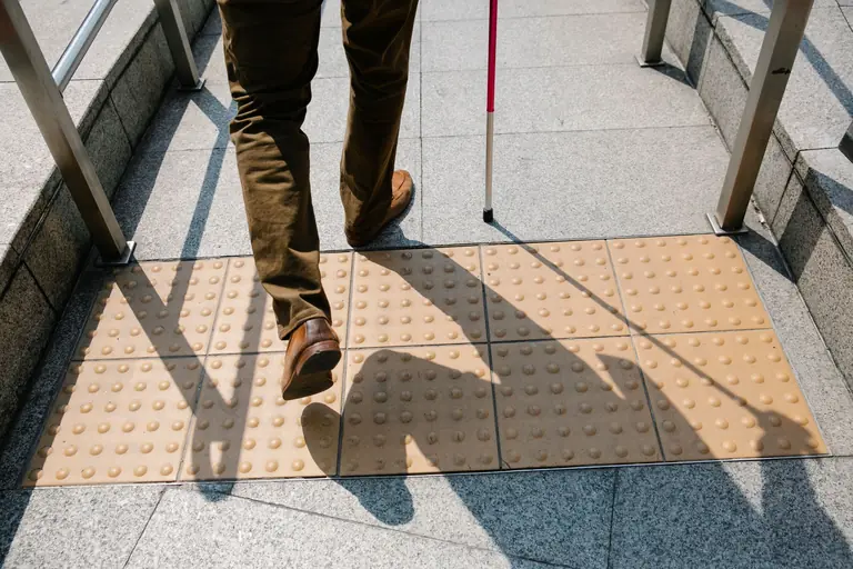 A person using a cane walks on tactile paving stones on a sunny day, emphasizing accessibility.