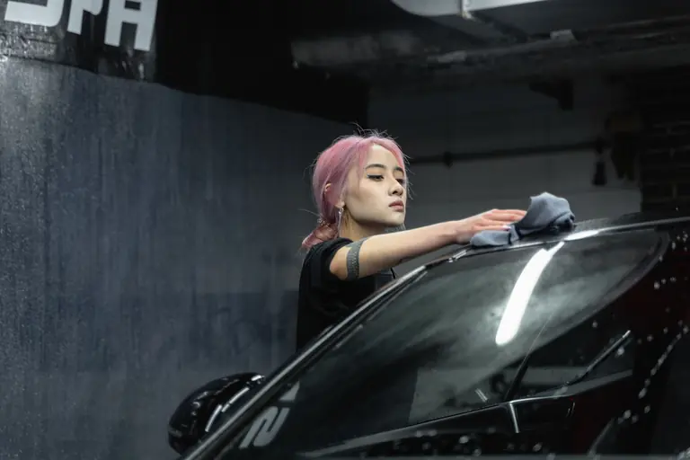 A young woman with pink hair wipes a car windshield with a microfiber cloth indoors.
