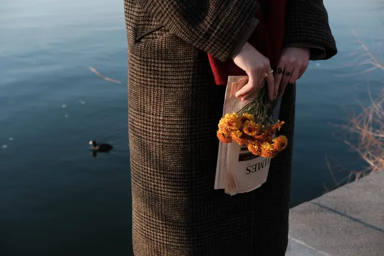 A woman in a coat holds yellow flowers and a newspaper by a calm lake, capturing a serene moment.