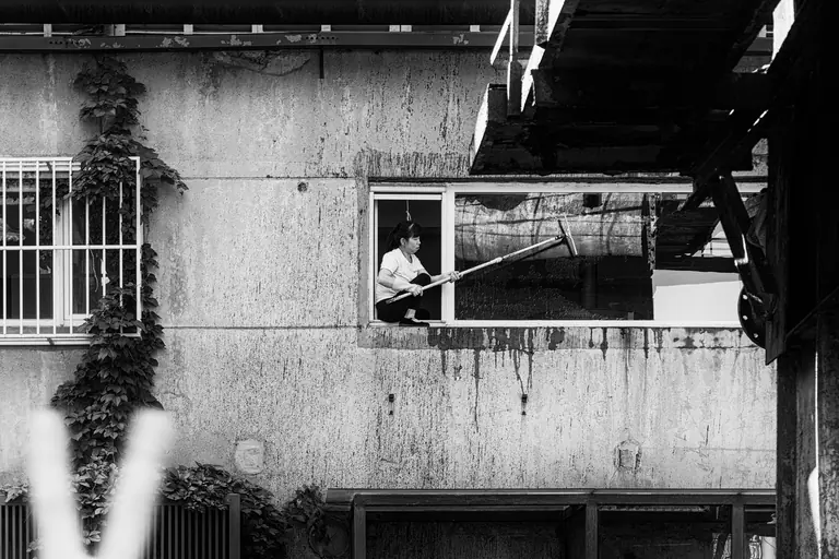 A woman cleaning a window in an urban setting, captured in black and white.